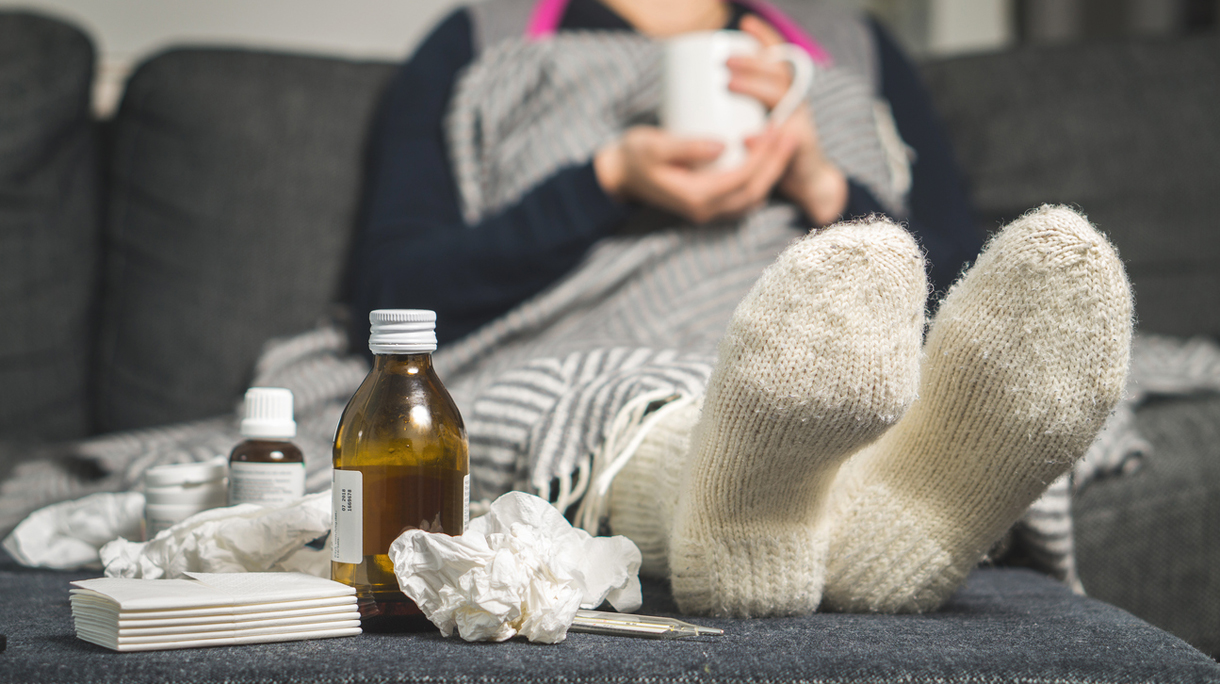 Cold medicine and sick woman drinking hot beverage to get well from flu, fever and virus. Dirty paper towels and tissues on table. Ill person wearing warm woolen stocking socks in winter.