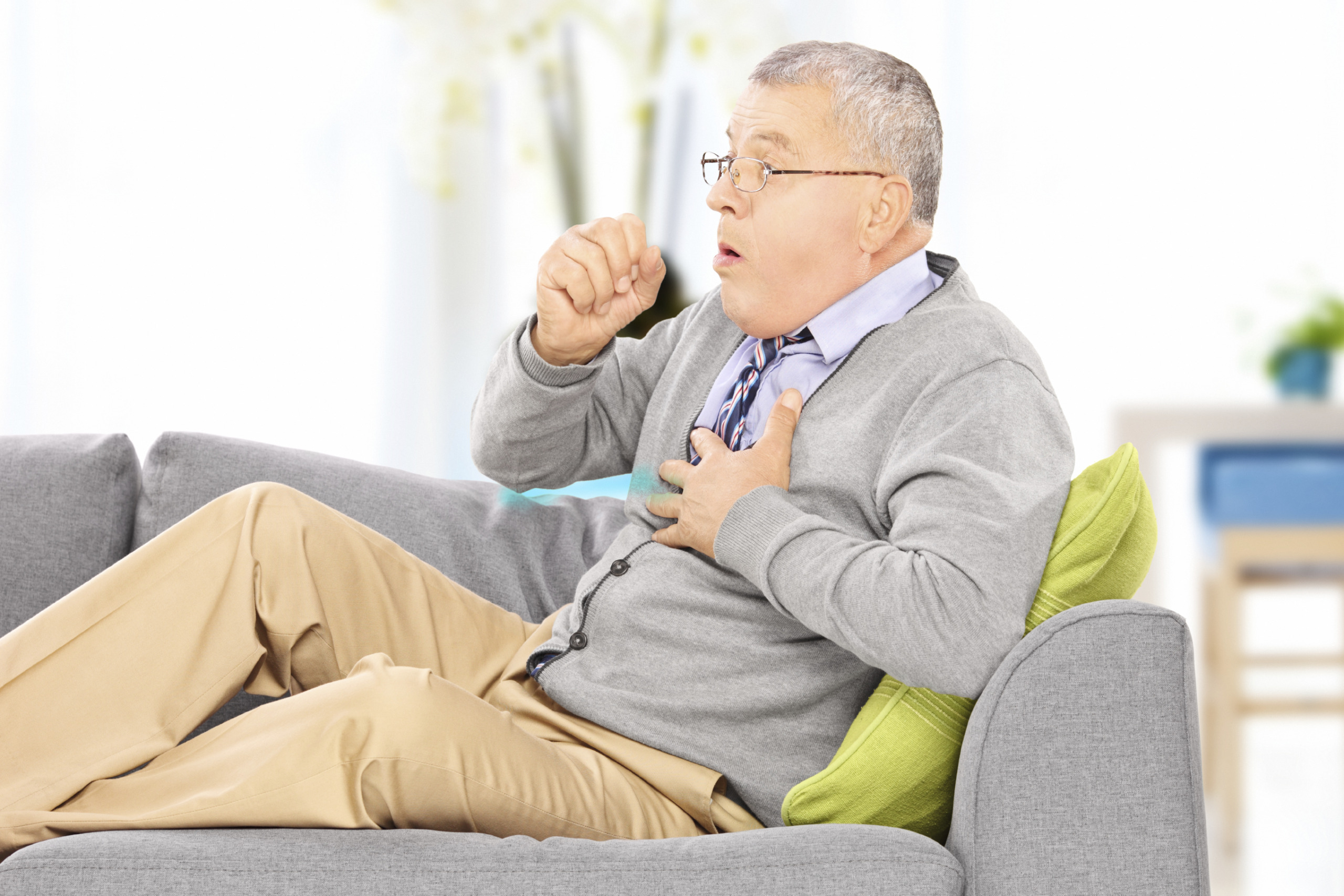 Mature man seated on a sofa coughing at home