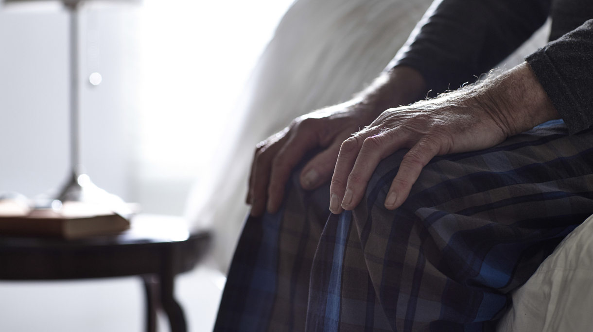 Senior man sitting on bed with his hands on knees