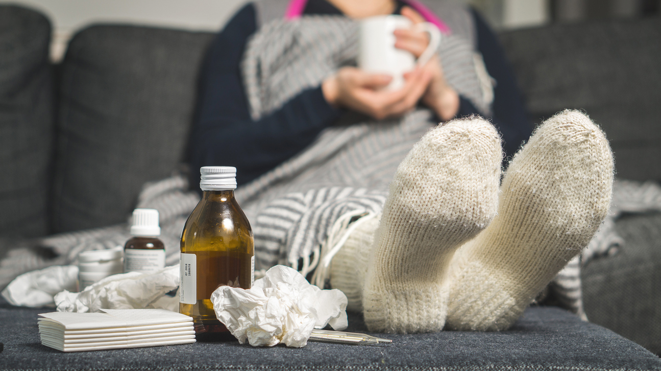 Cold medicine and sick woman drinking hot beverage to get well from flu, fever and virus. Dirty paper towels and tissues on table. Ill person wearing warm woolen stocking socks in winter.