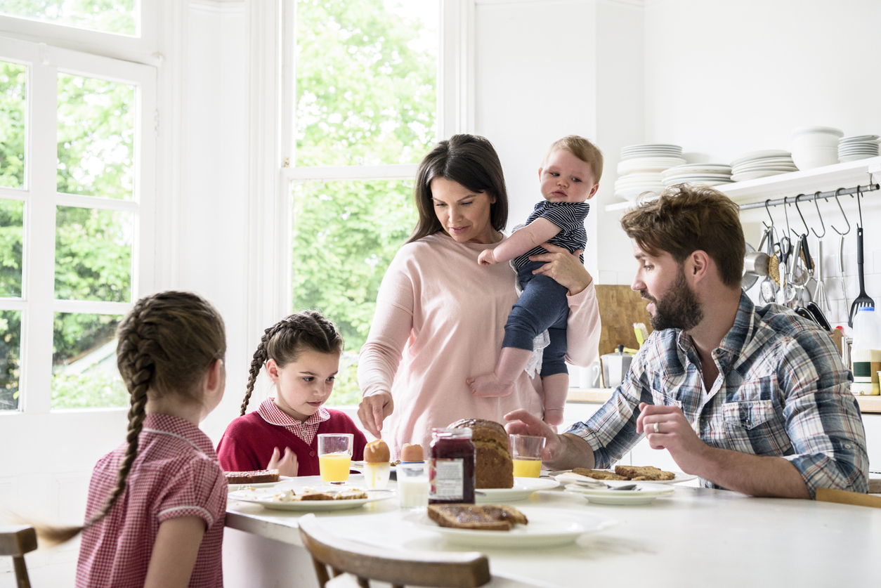 Family sitting at breakfast table, mother holding baby boy, father and girls eating food