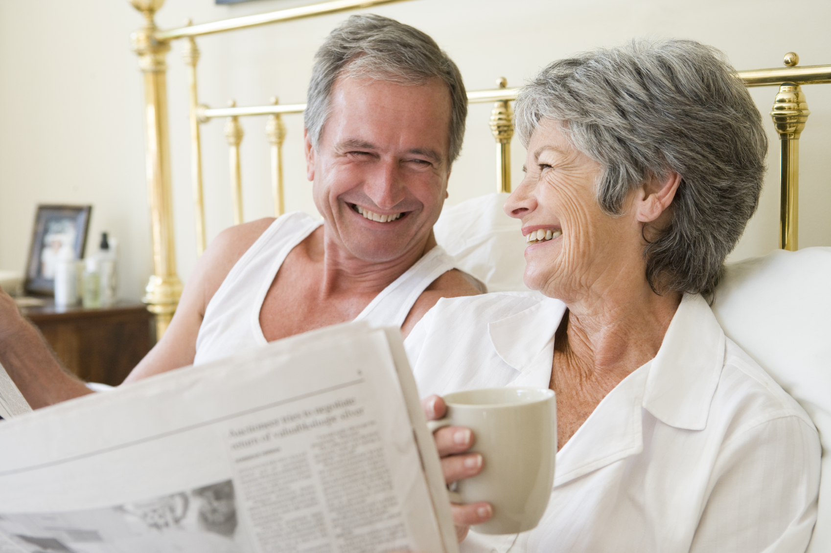 Couple in bedroom with coffee and newspapers smiling