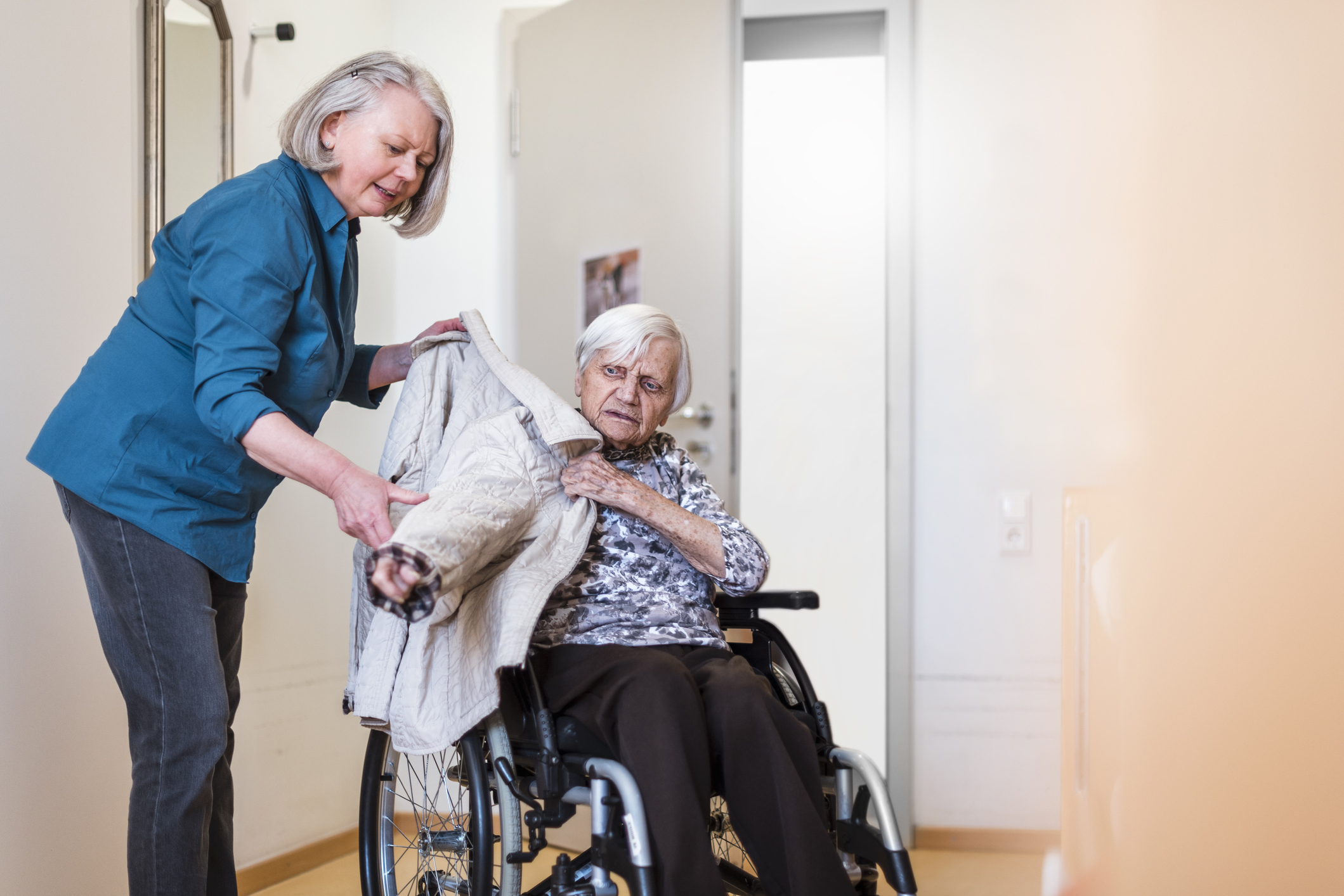 Woman taking care of old woman in wheelchair putting her jacket on