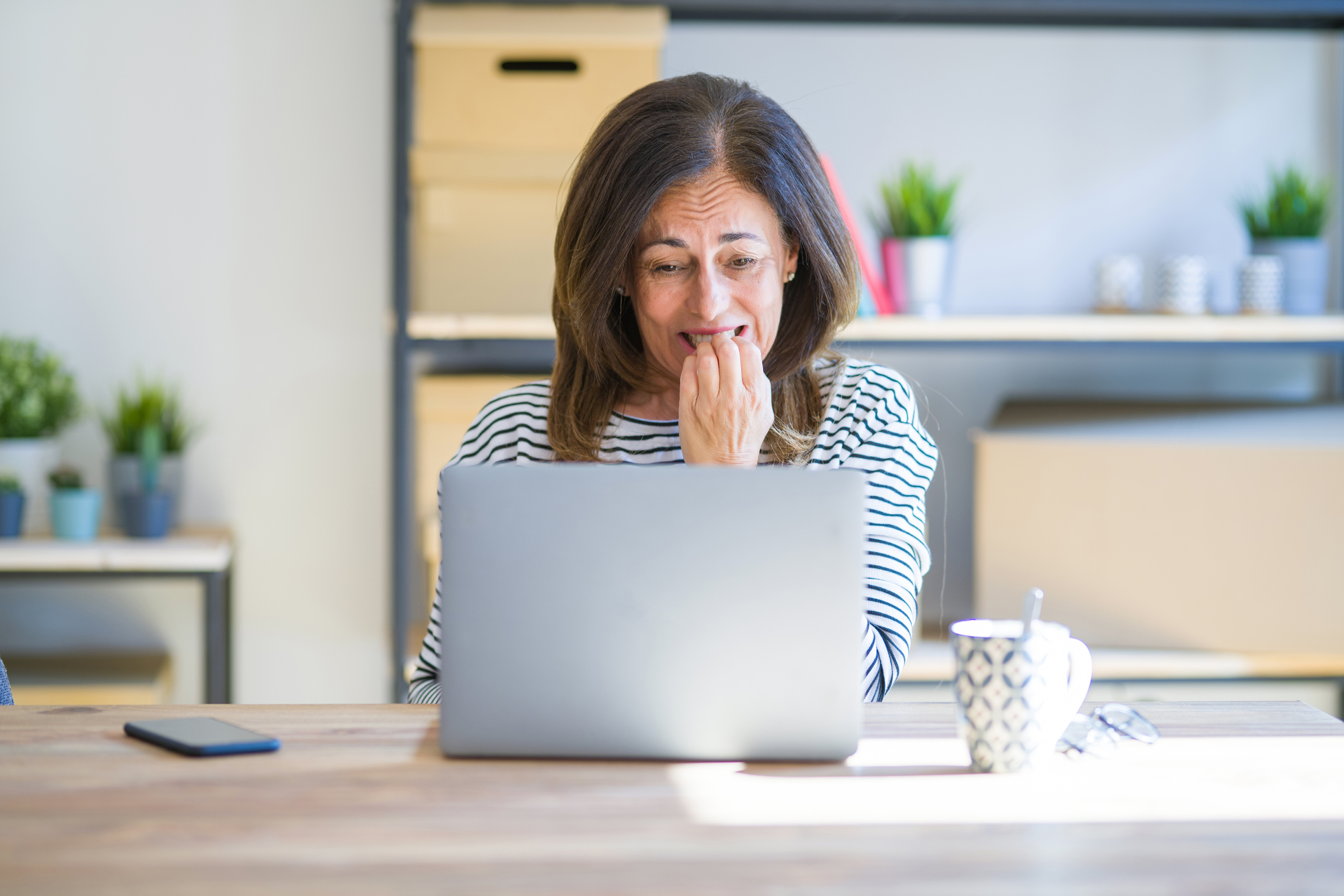 Middle age senior woman sitting at the table at home working using computer laptop looking stressed and nervous with hands on mouth biting nails. Anxiety problem.