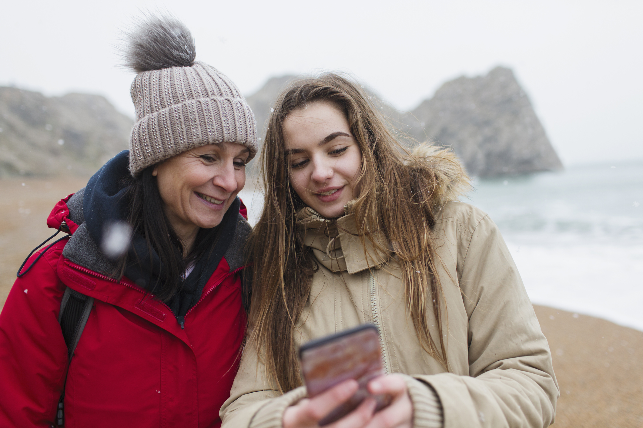 Mother and daughter in warm clothing using smart phone on snowy winter beach