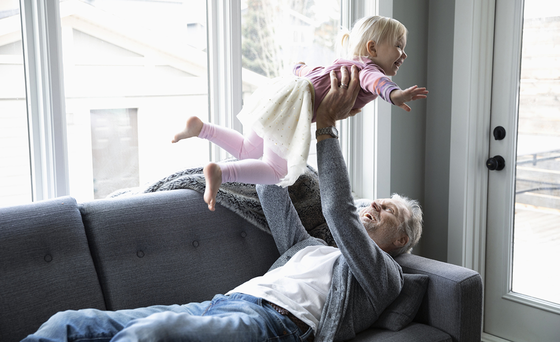 Playful grandfather lifting toddler granddaughter overhead on living room sofa