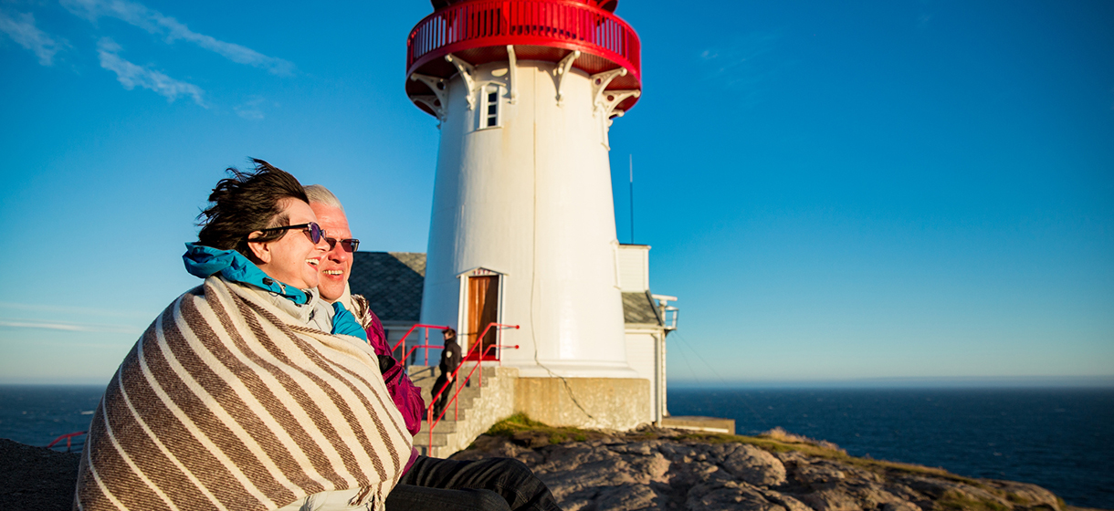 Loving mature couple hiking, sitting on windy top of rock with lighthouse.