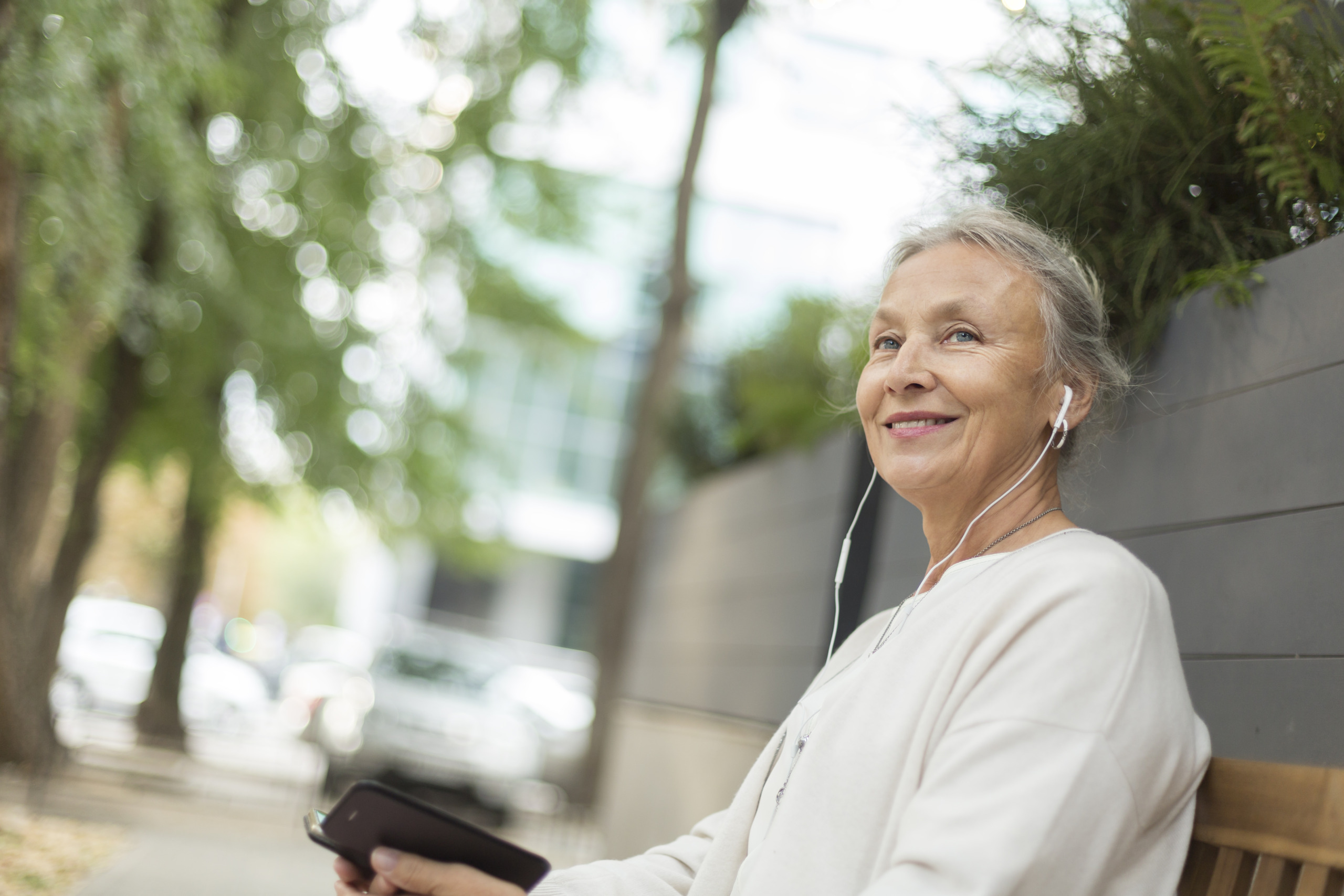 Smiling senior woman sitting on a bench outdoors with cell phone and earphones