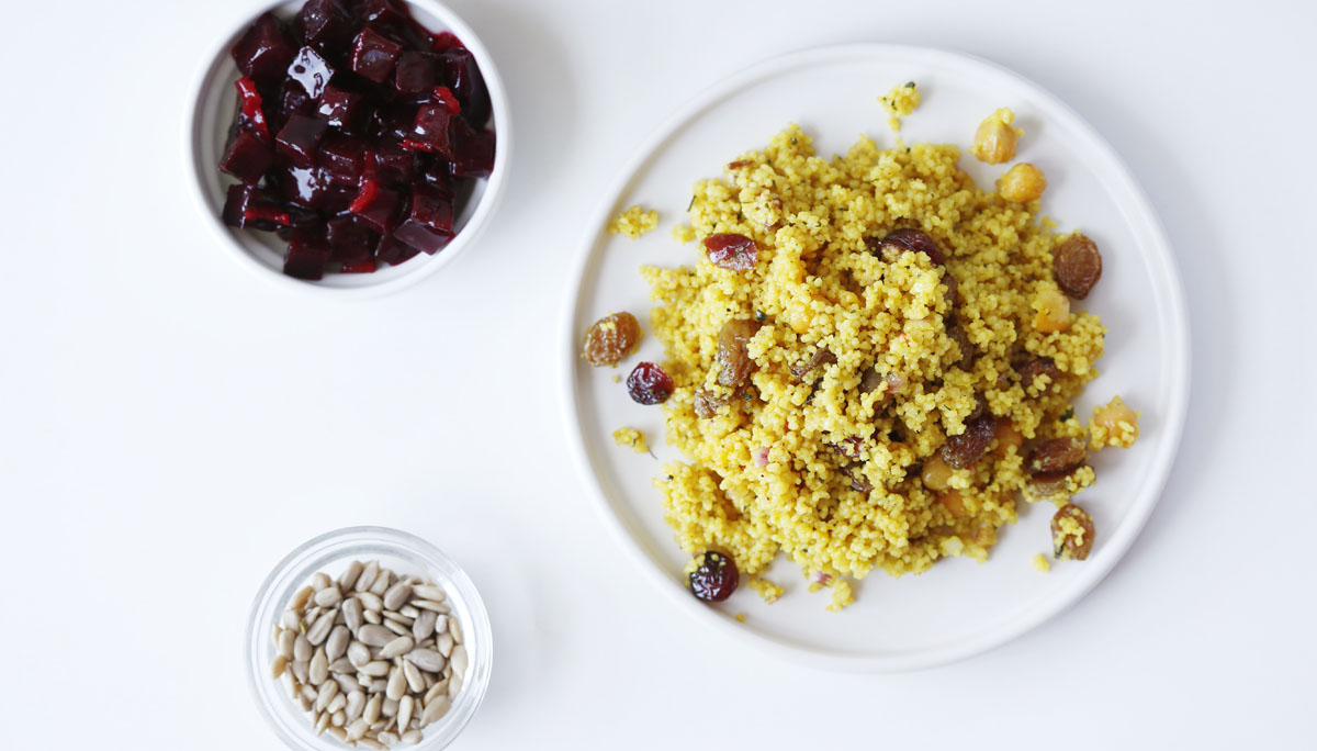 Overhead view of couscous served in plate with beetroot salad and seeds in bowls on white background
