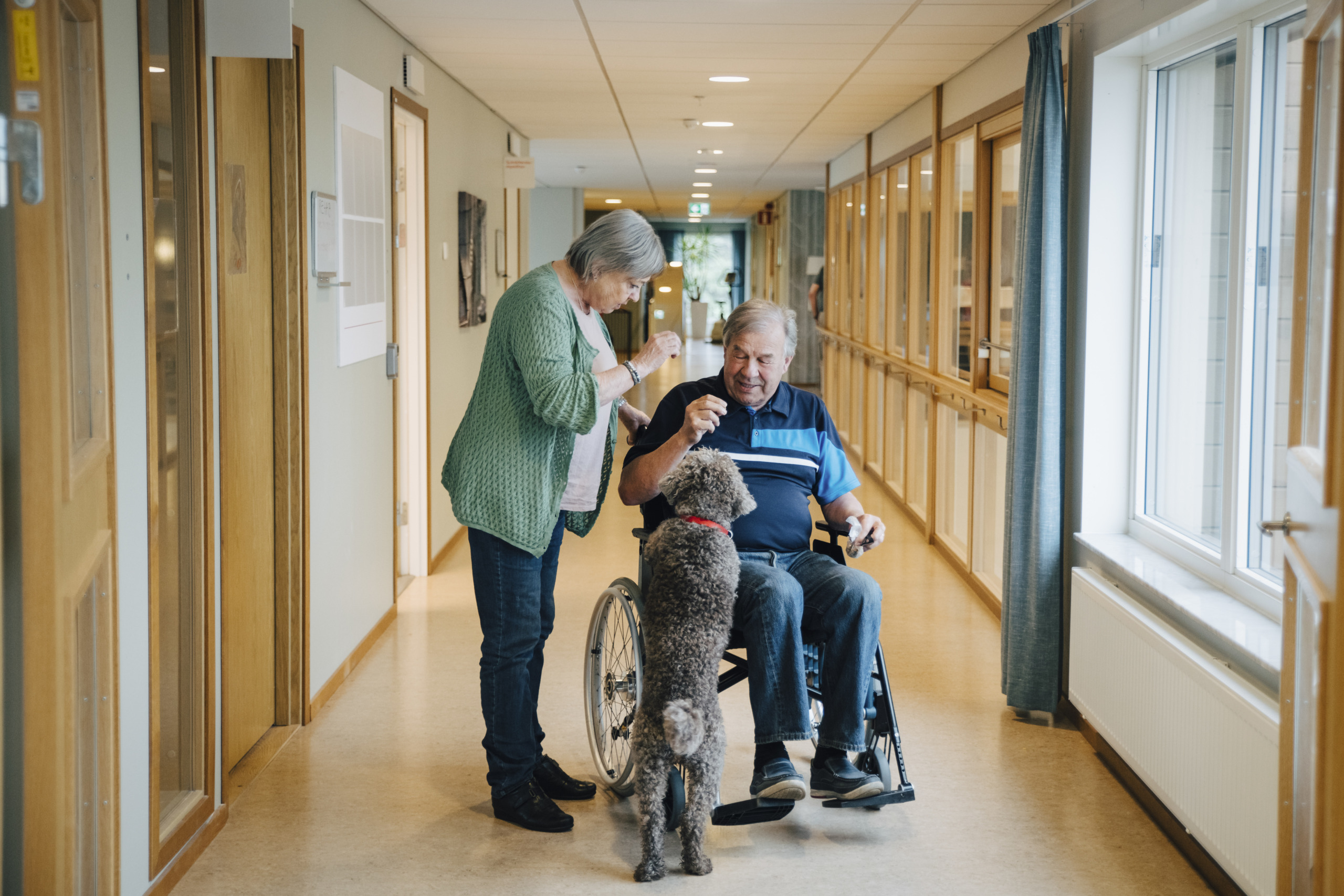 Senior woman standing by disabled man on wheelchair with dog in alley at retirement home