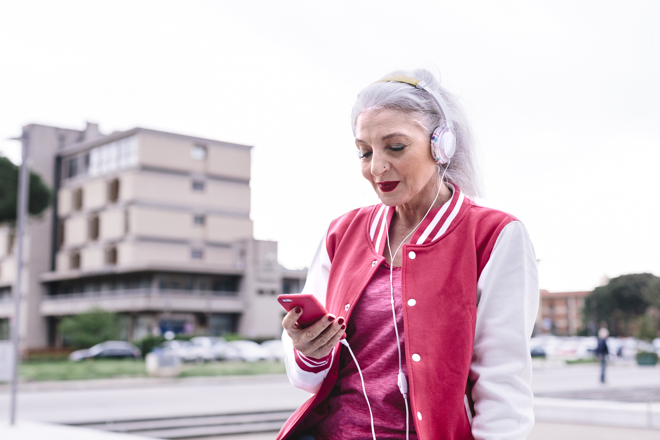 Mature woman in baseball jacket listening to headphones and looking at smartphone
