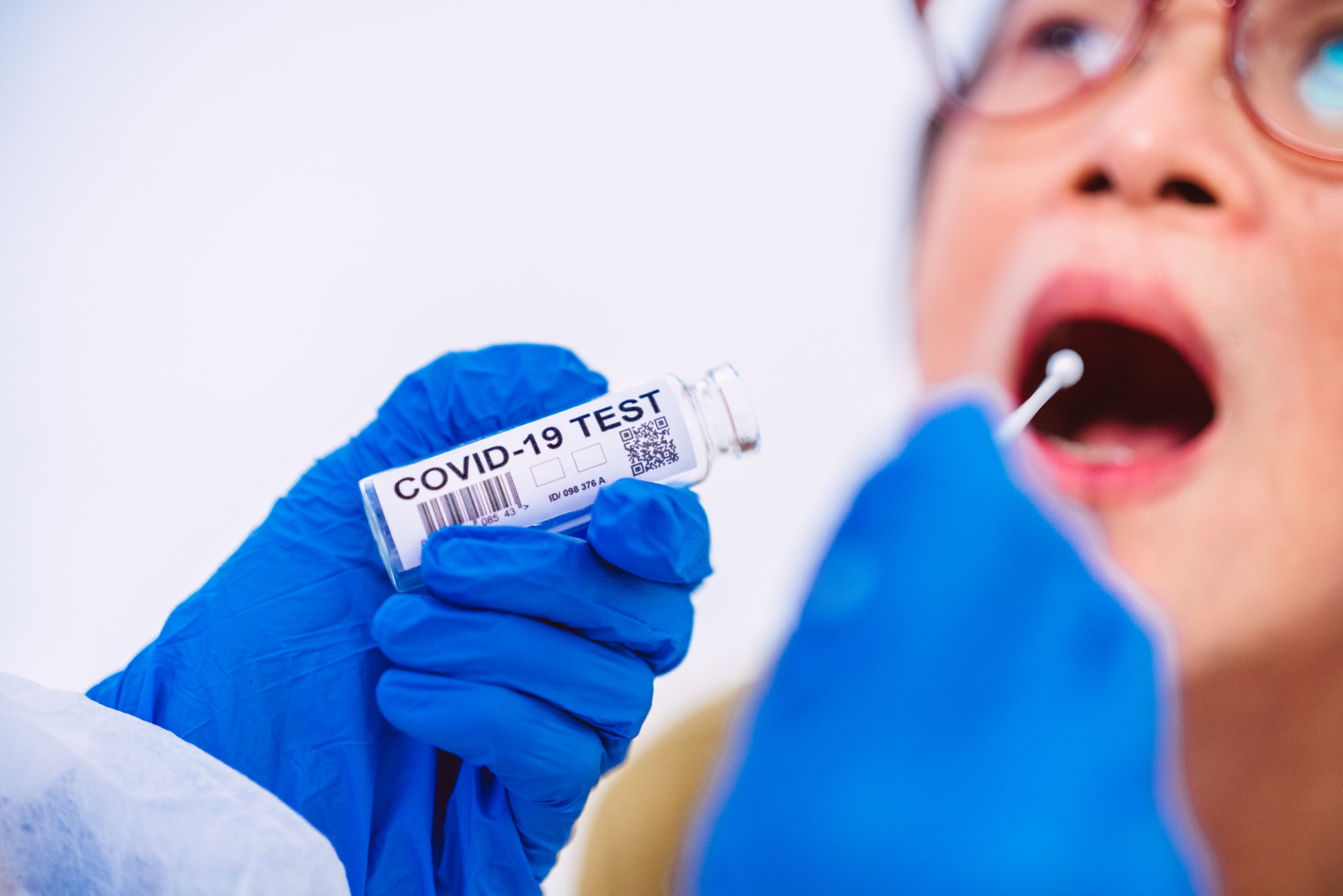 Doctor’s hands in protective gloves & workwear putting COVID-19 test swab into senior woman’s mouth
