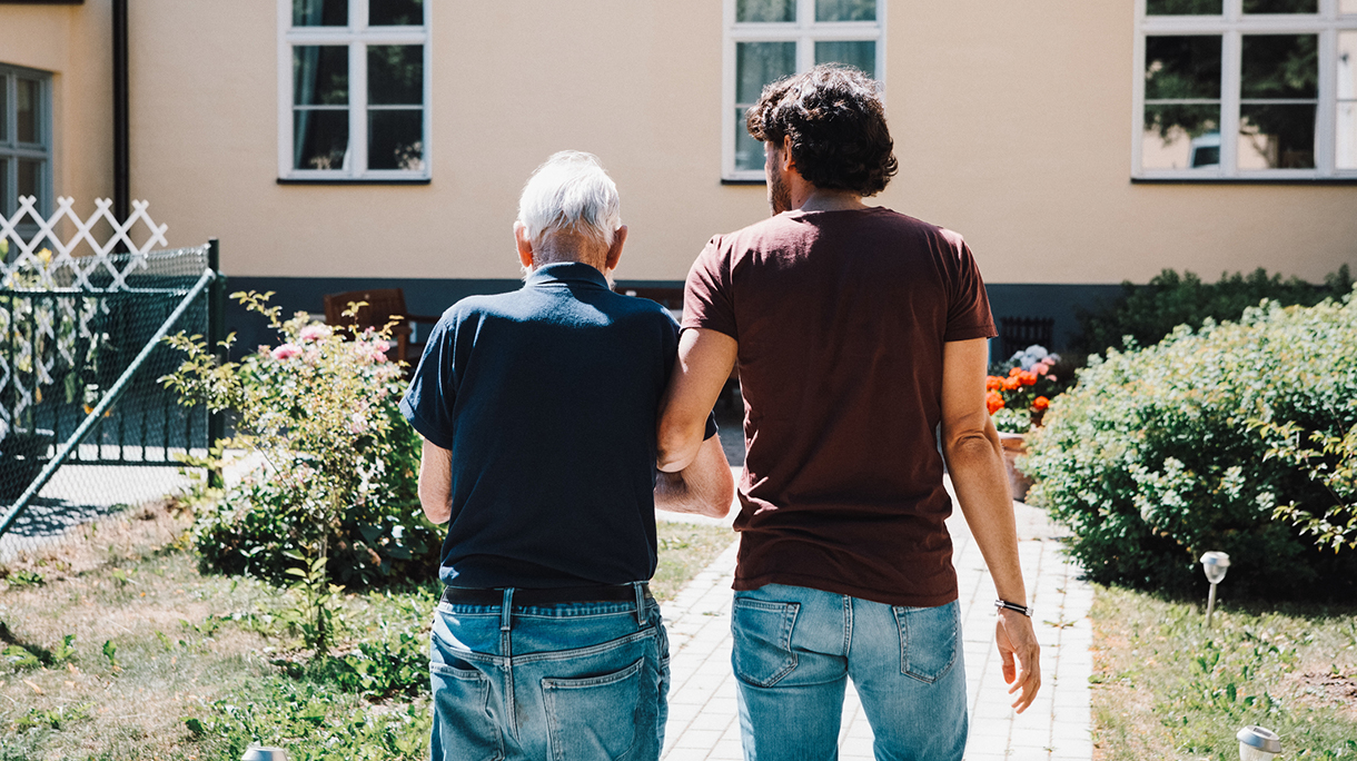 Rear view of male caretaker walking arm in arm with senior man at nursing home