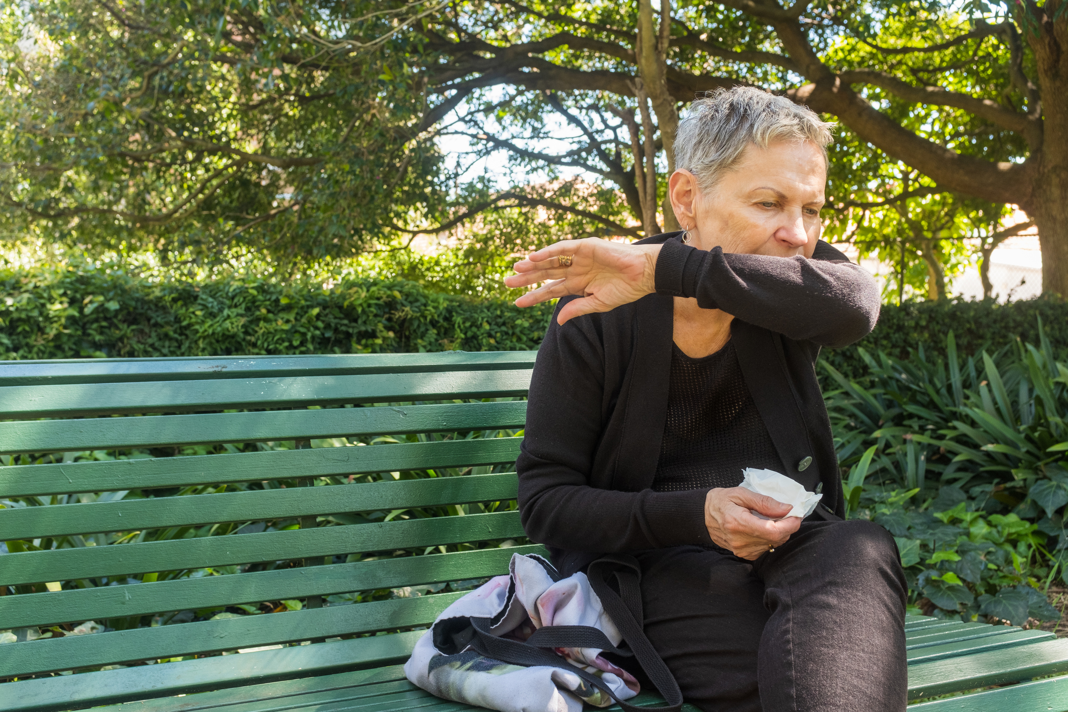 Elderly Woman Sitting On Bench Outdoors Coughing Into Elbow And Holding Tissue – Coronavirus Concept