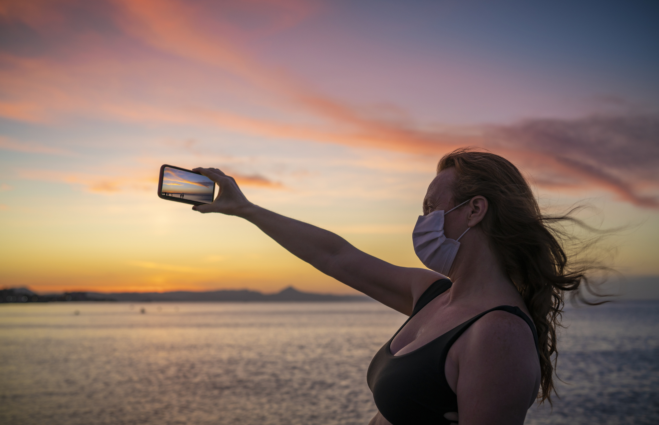 Tourist adult 50s  woman in pandemic  face mask at sunset beach taking photo