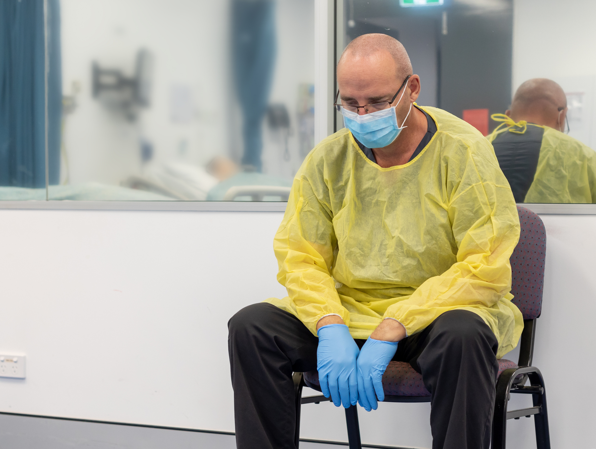 Male Health care sitting in a chair beside a closed hospital room looking exhausted