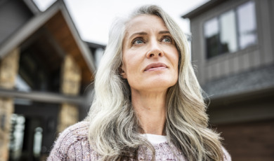 Knappe oudere vrouw met prachtig golvend grijs haar staat voor haar huis en kijkt bedachtzaam