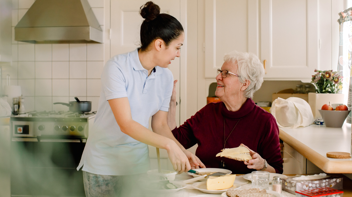 Smiling retired elderly woman talking with female caregiver in kitchen at nursing home