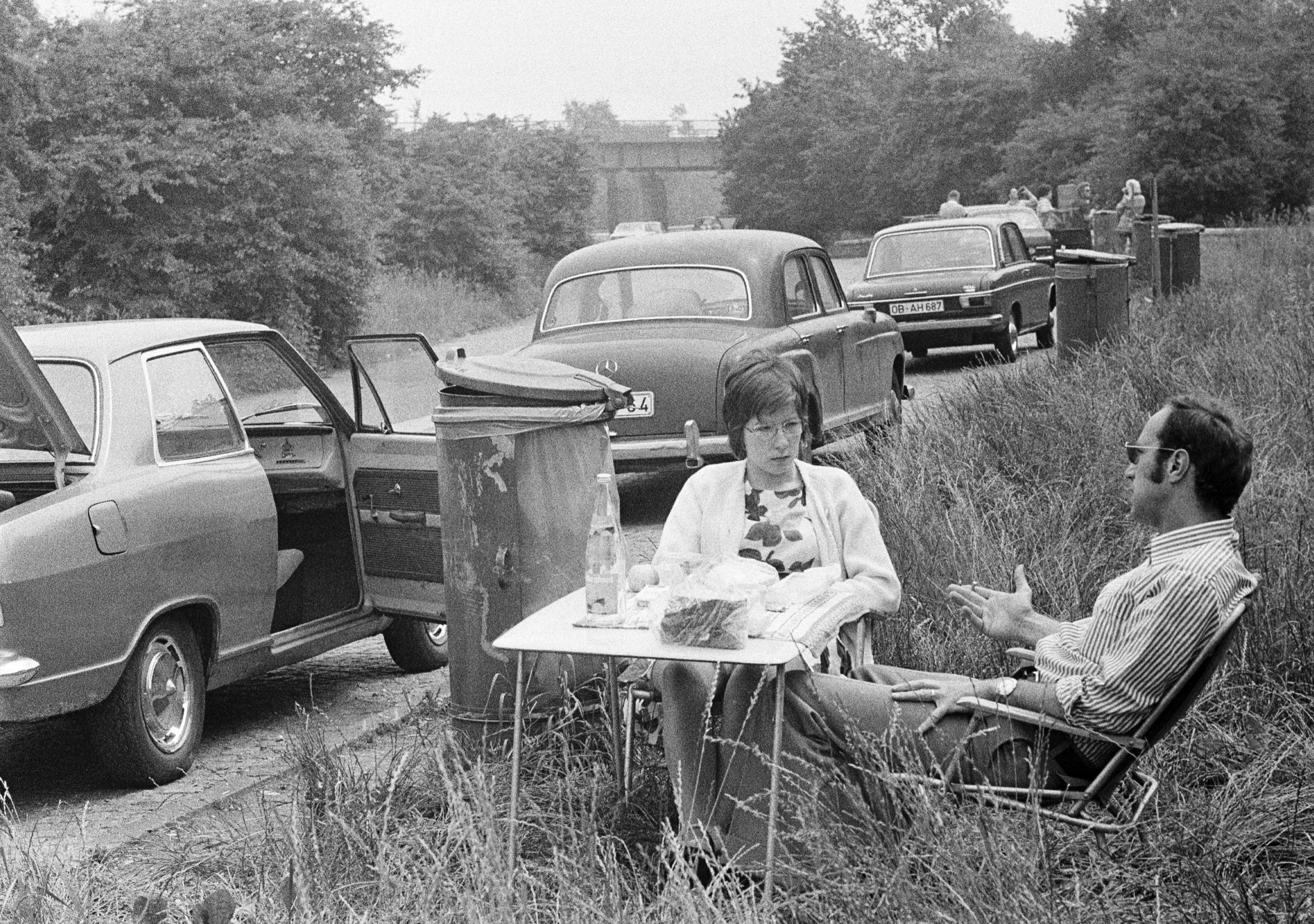 people, freetime, young couple takes a rest from motoring in a rest area, aged 25 to 35 years, picnic with folding chairs and a folding table, in the background parking cars and a dustbin – 30.06.1973