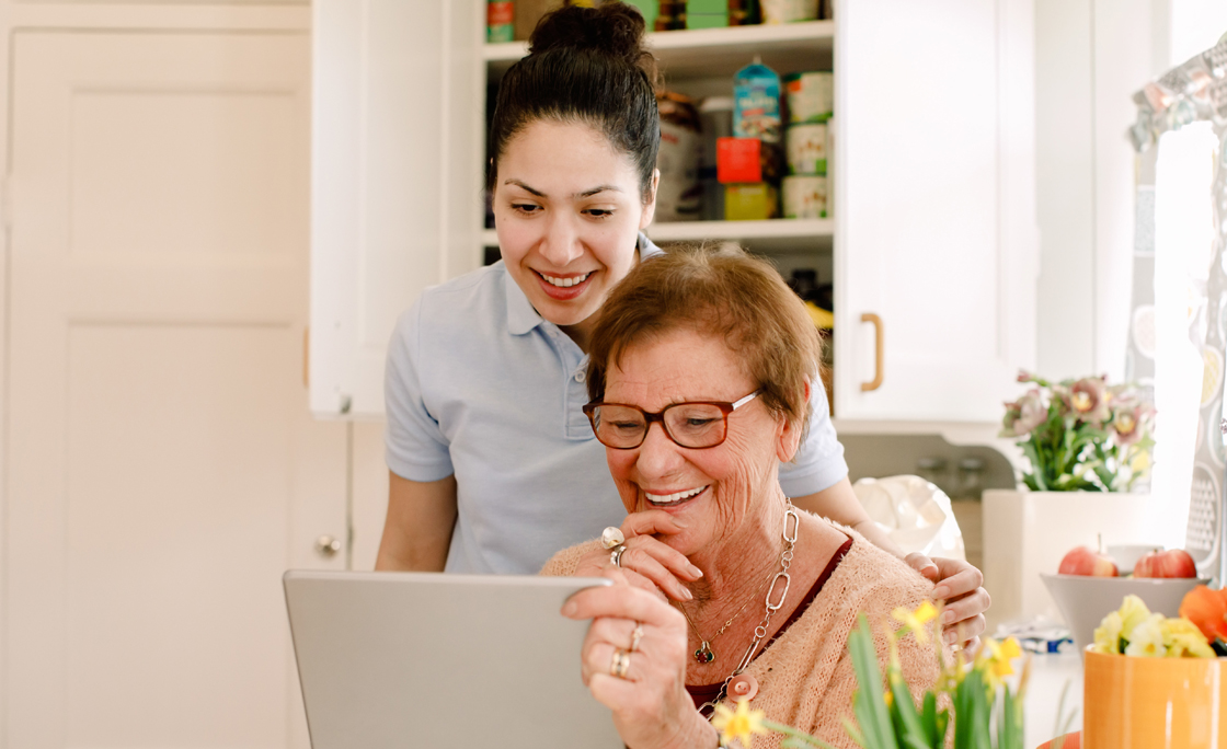 Smiling elderly woman and young female caregiver looking at digital tablet in nursing home kitchen