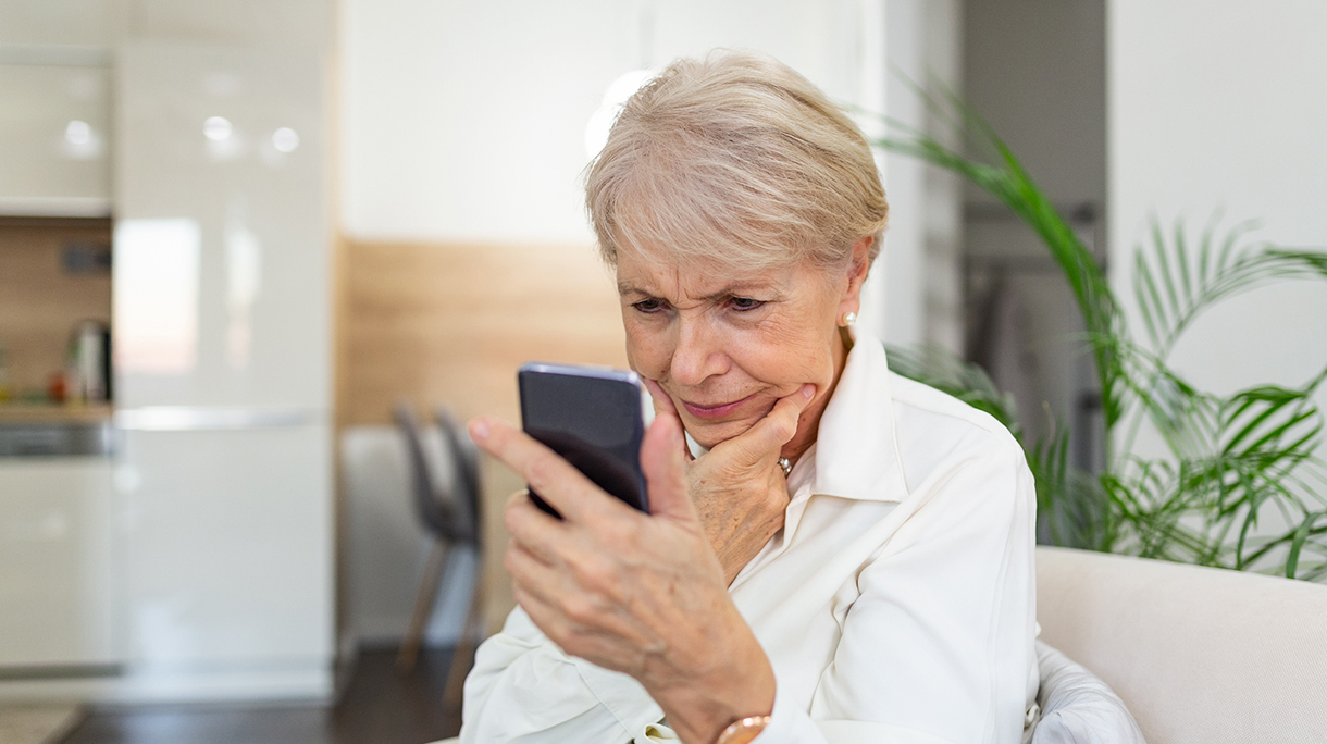 Confused senior woman having trouble using mobile phone at home. Old woman with white hair sitting on sofa and trying to messaging with smartphone.