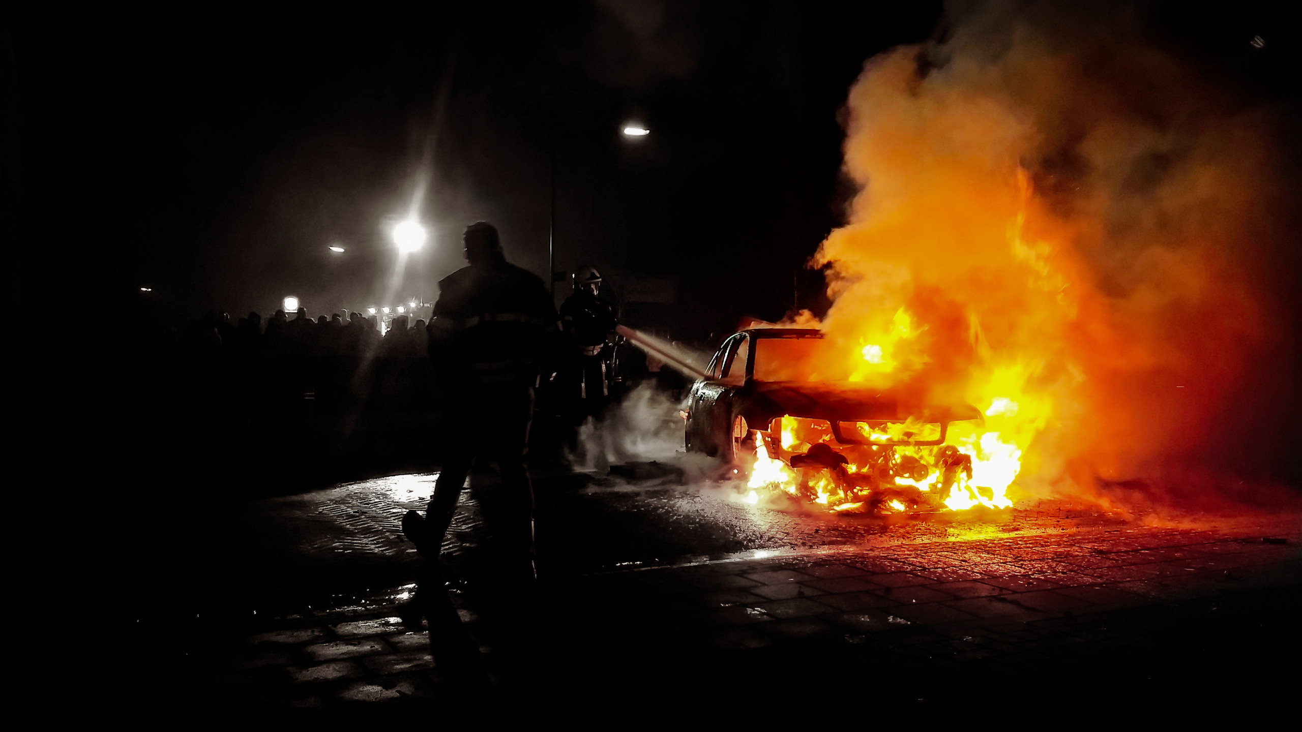 Firefighters Standing By Burning Car On Street At Night