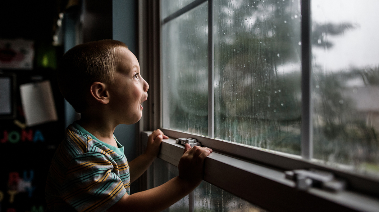excited boy looking out a window with raindrops at a stormy sky