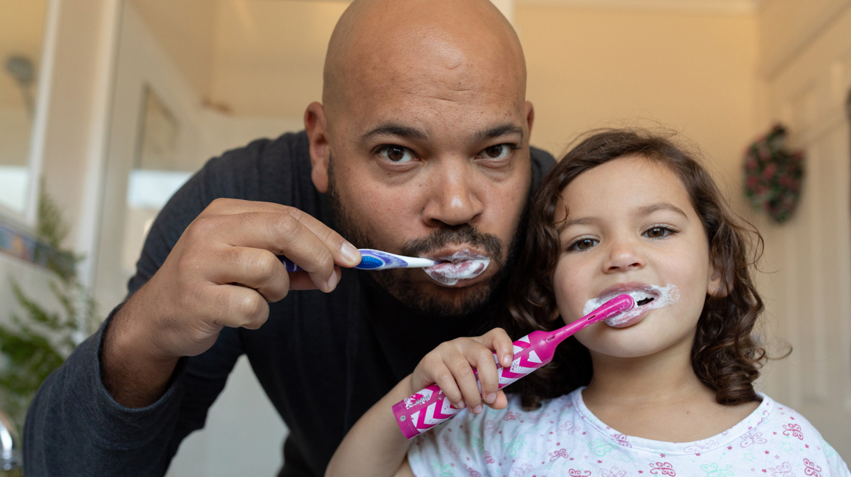 Father and daughter brushing their teeth and looking into camera