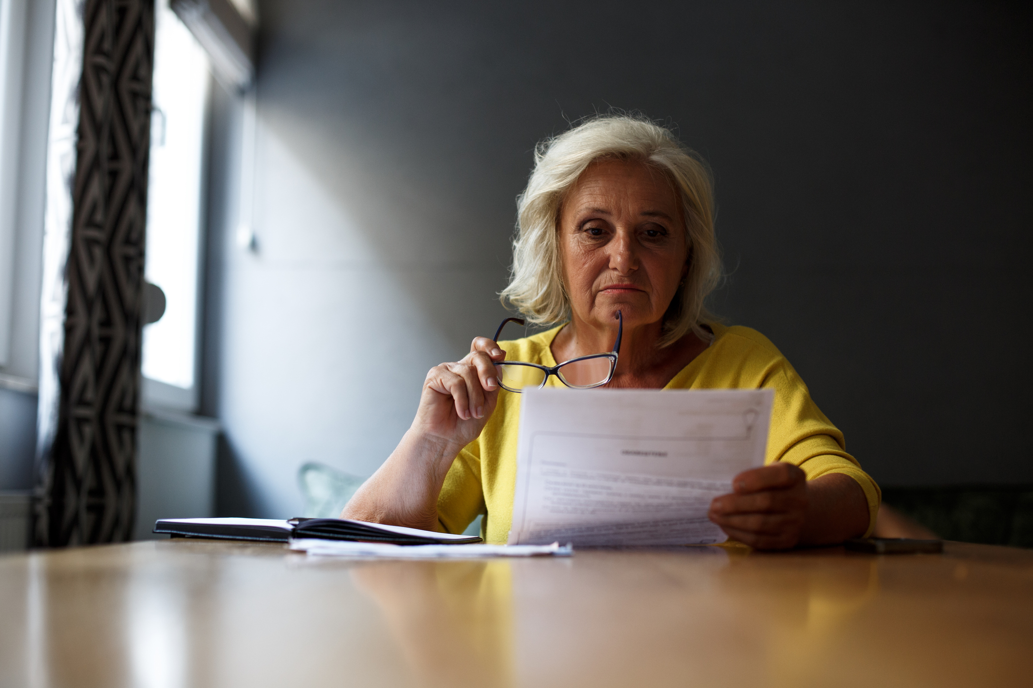 Senior lady in yellow blouse is sitting at the table and doing paperwork.
