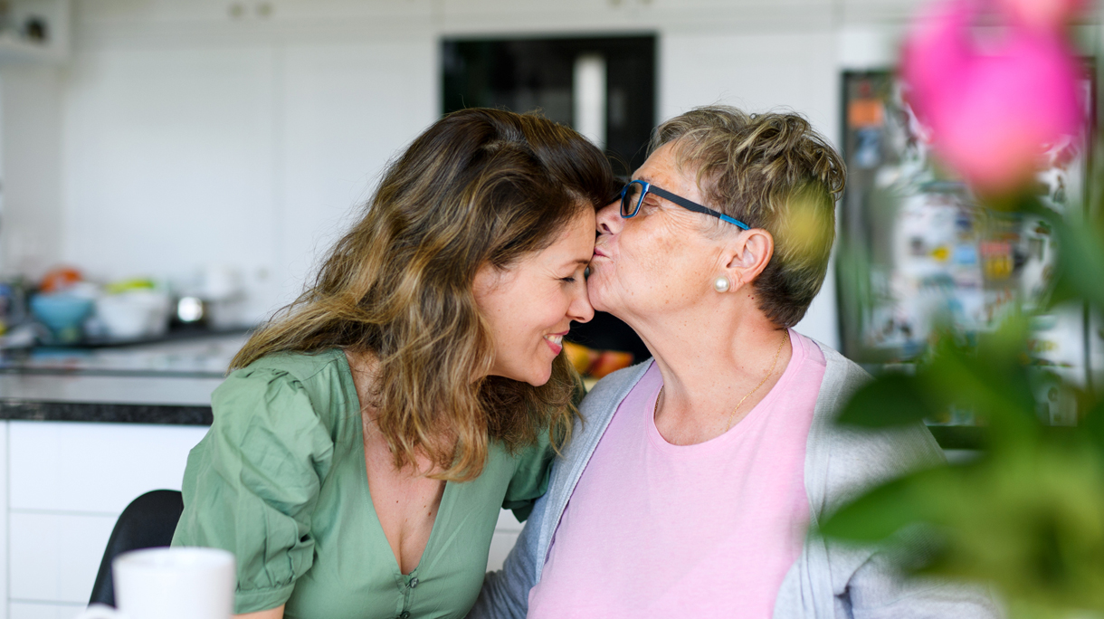 Senior mother kissing adult daughter on forehead indoors at home.