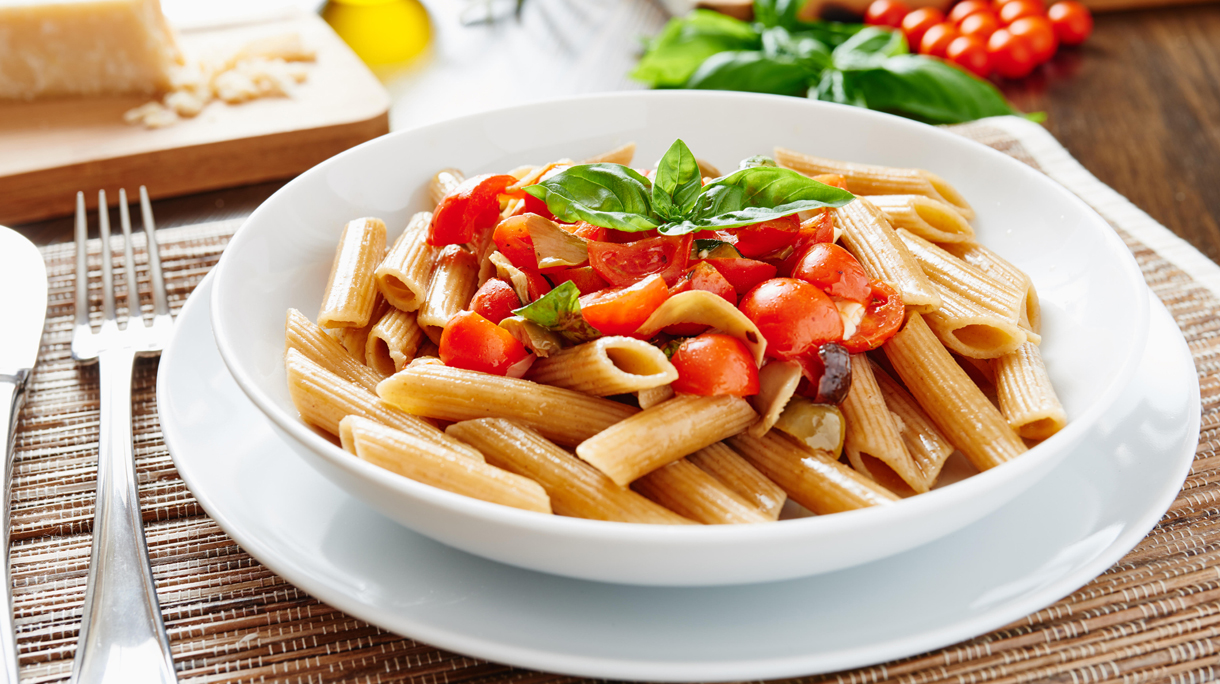 Close-up of pasta penne with cherry tomatoes on white plate surrounded by ingredients – vegan food