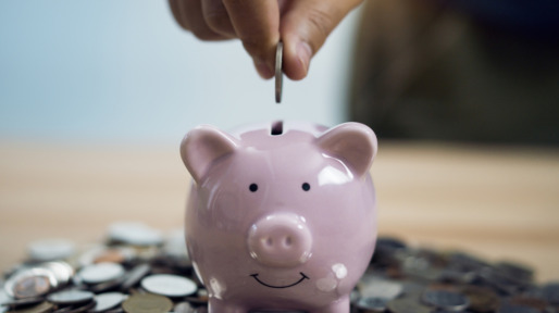 Person Putting Coin In Piggy Bank At Table