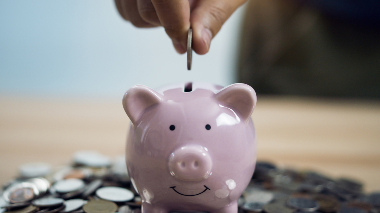 Person Putting Coin In Piggy Bank At Table