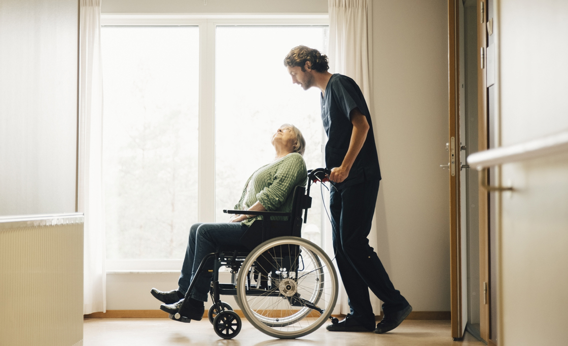 Full length side view of male nurse pushing disabled senior woman on wheelchair in alley at retirement home