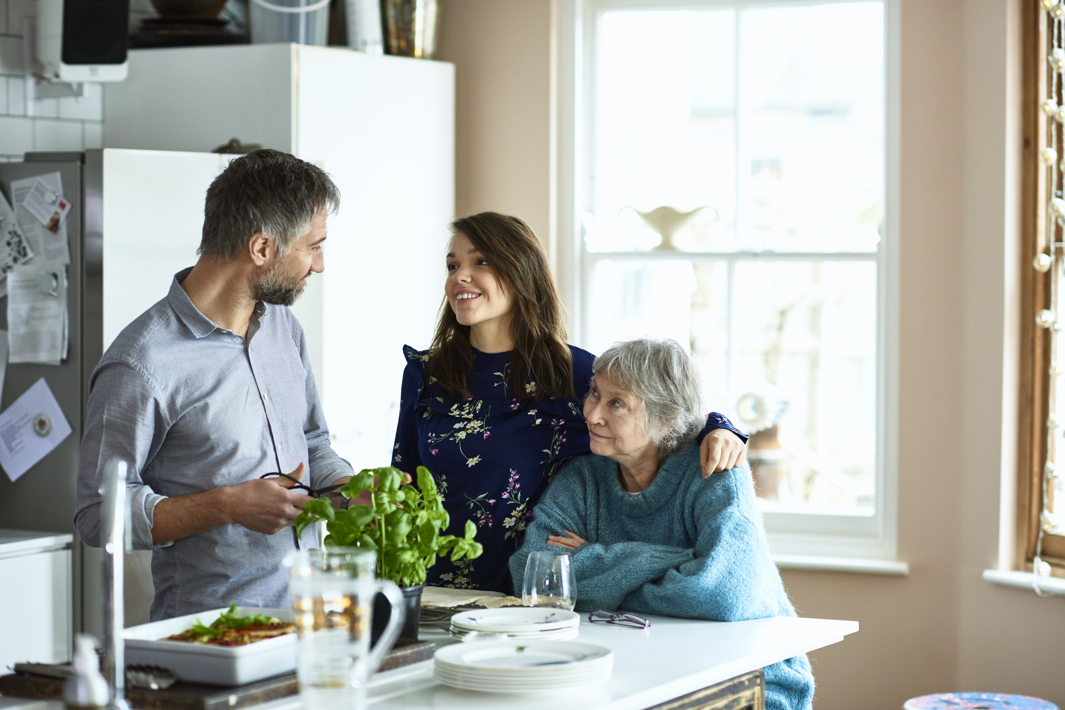 Woman with arm around mature woman looking towards man in kitchen