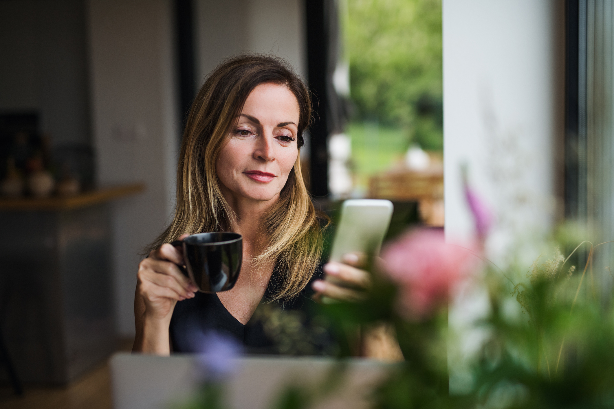 Front view portrait of mature woman with laptop sitting indoors at the table, using smartphone.