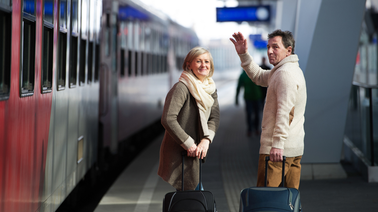 Senior couple on train station pulling trolley luggage, waving.
