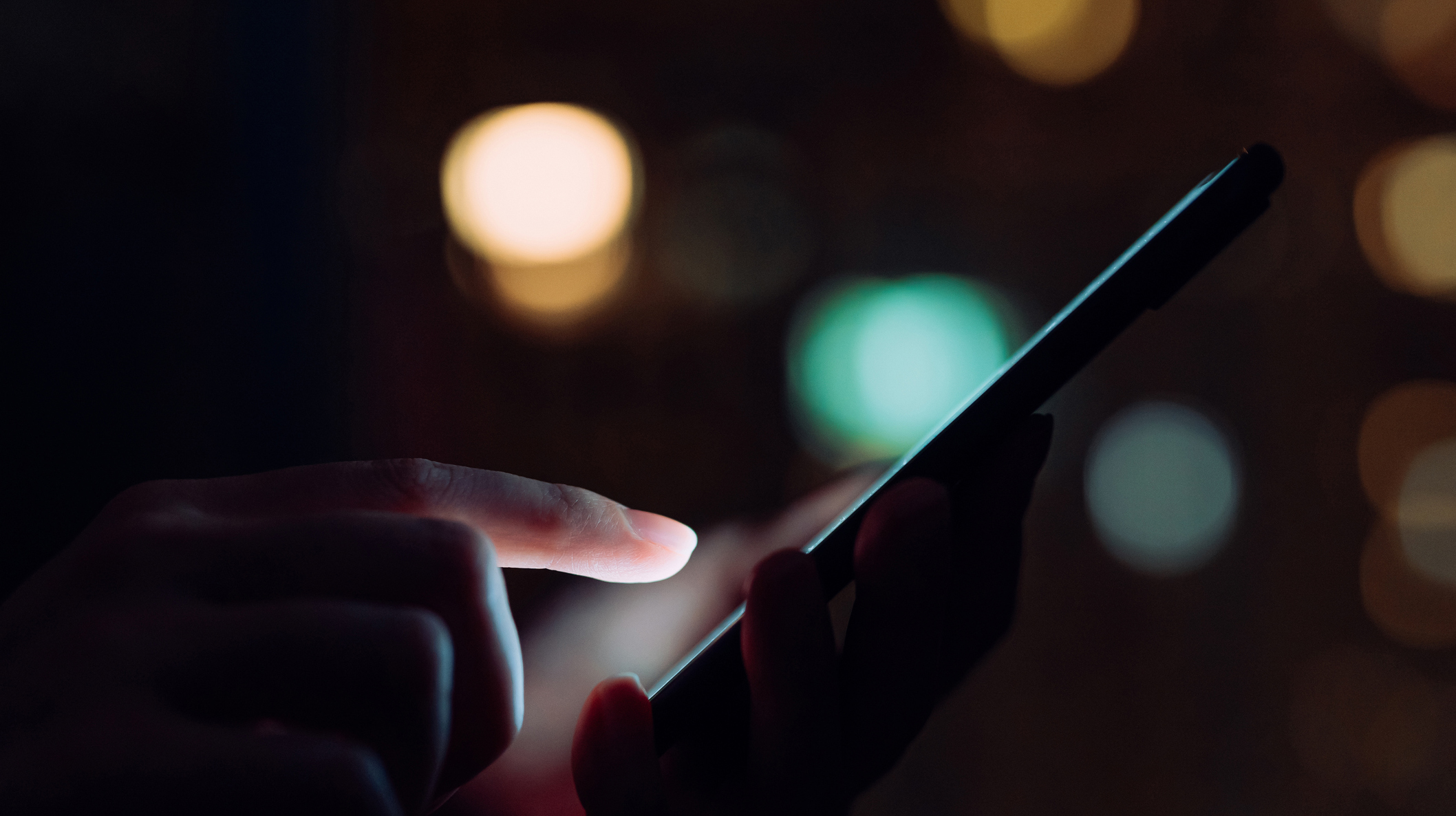 Close up of woman’s hand using smartphone in the dark, against illuminated city light bokeh