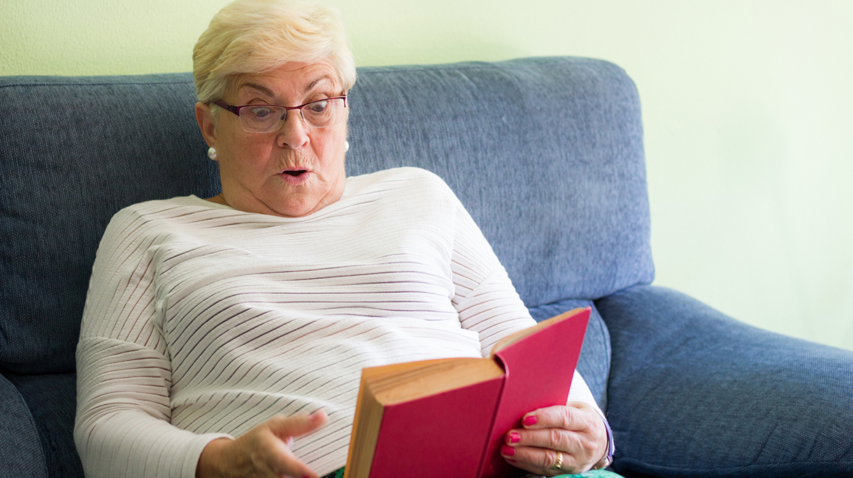 Scared senior woman with eyeglasses reading red book sitting on sofa