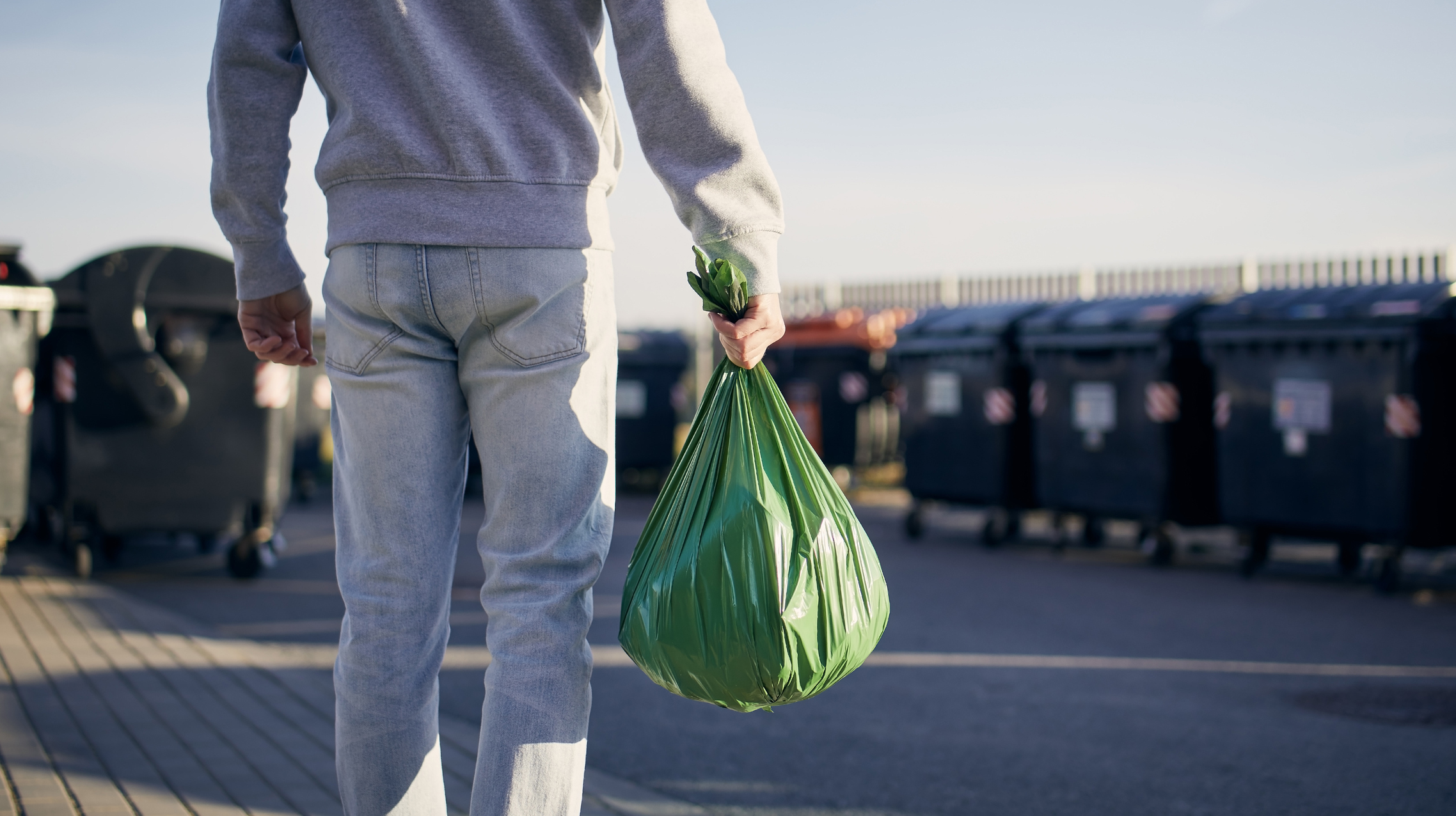 Man Walking With Rubbish. Rear View Of Person Carrying Plastic Bag Against Garbage Cans On Street.