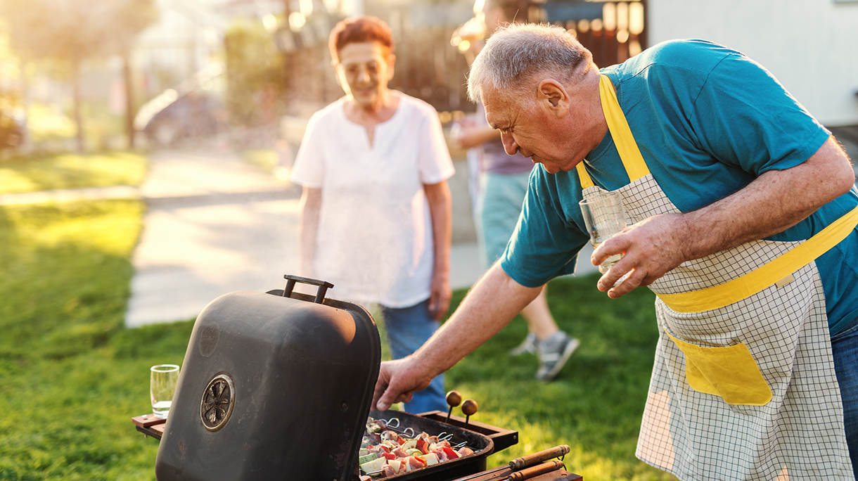 Grandfather with apron checking on grill and holding glass of beer while standing in the backyard. In background his wife. Family gathering concept.