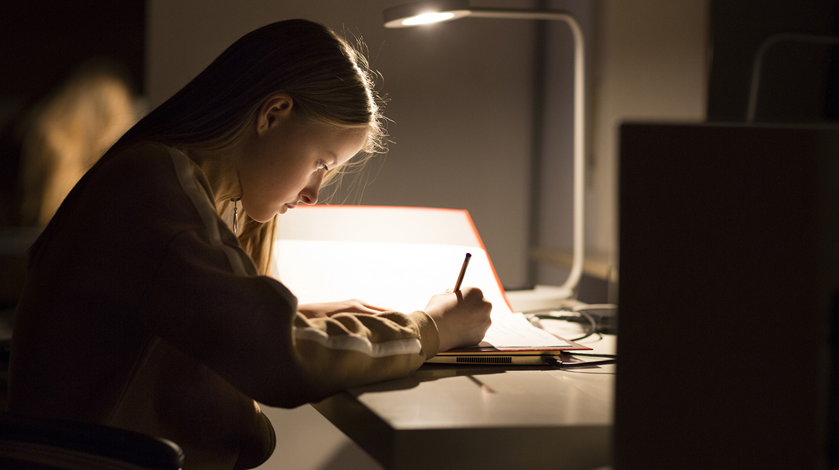 Girl (12-13) doing homework at night on a desk lit by a lamp