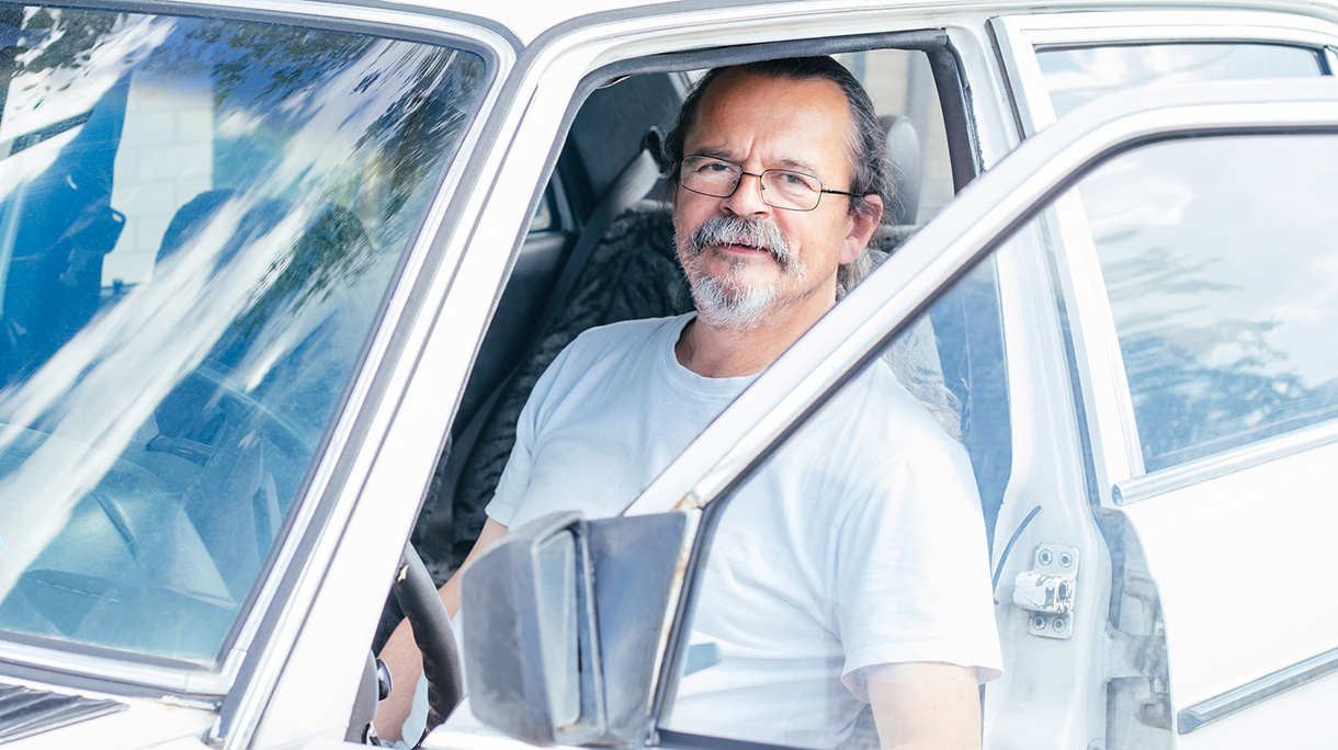 Senior man with gray mustache wearing glasses and white t-shirt sits in white car with open door smiling