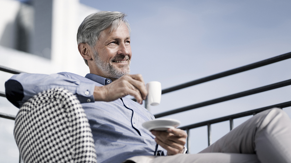 Smiling grey-haired man sitting on balcony and drinking coffee