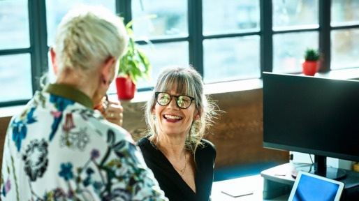 Cheerful mature woman at desk smiling towards senior female colleague