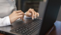 Beautiful young woman in white t-shirt is working on laptop and smiling while sitting outdoors in cafe. Young female using laptop for work. Female freelancer working on laptop in an outdoor cafe.