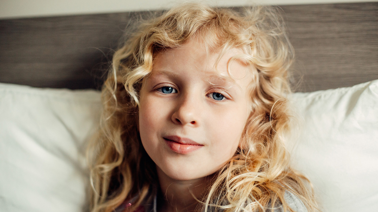 Pretty Beautiful Caucasian Blonde Girl With Long Messy Hair And Blue Eyes Sitting On A Bed At Home
