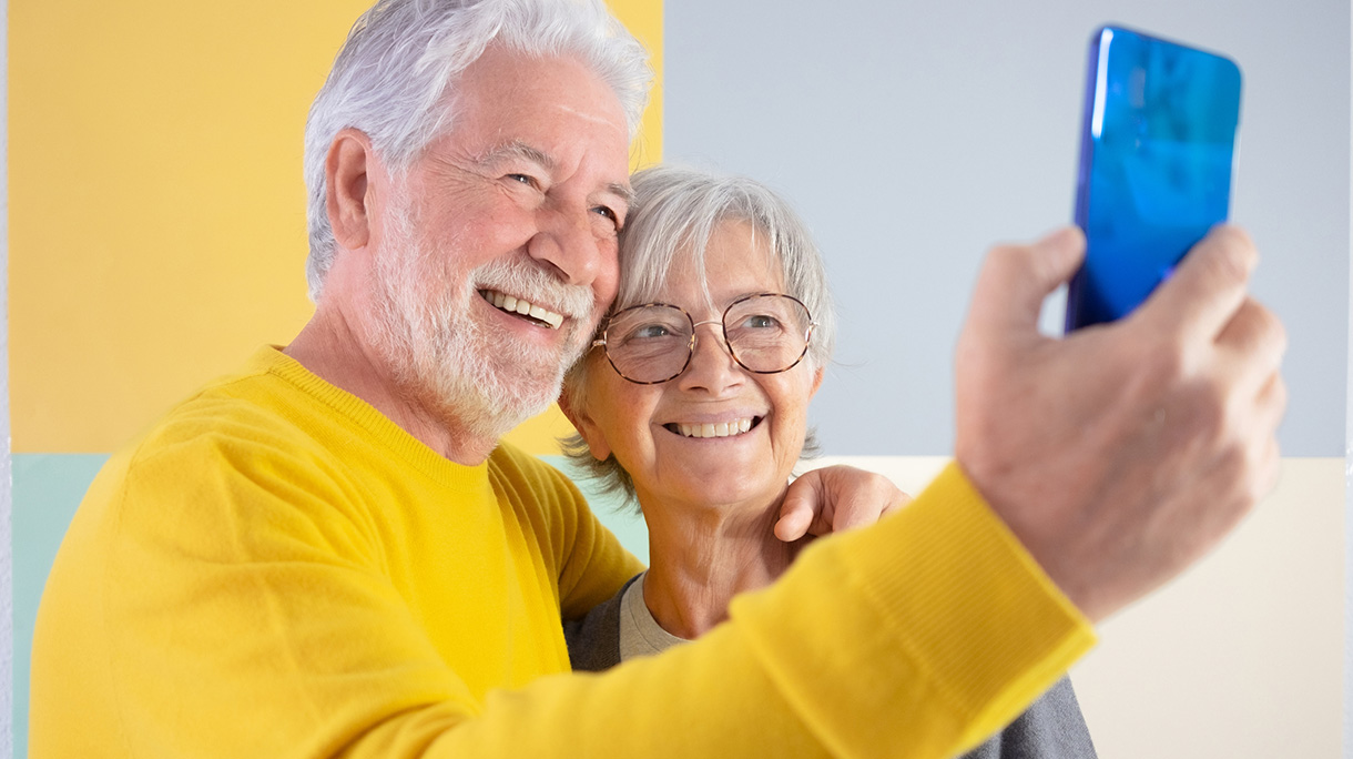 Happy caucasian beautiful senior couple standing over isolated colored background video calling with smart phone. Elderly white-haired people smiling carefree enjoying retirement