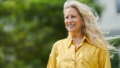 Vrouw met geel shirt en mooi blond haar kijkt lachend uit over haar tuin