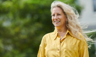 Vrouw met geel shirt en mooi blond haar kijkt lachend uit over haar tuin